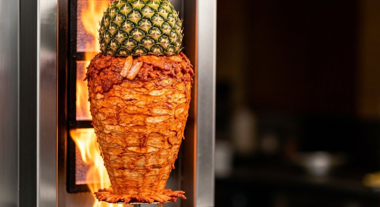 A rotating al pastor trompo charring beside an open flame